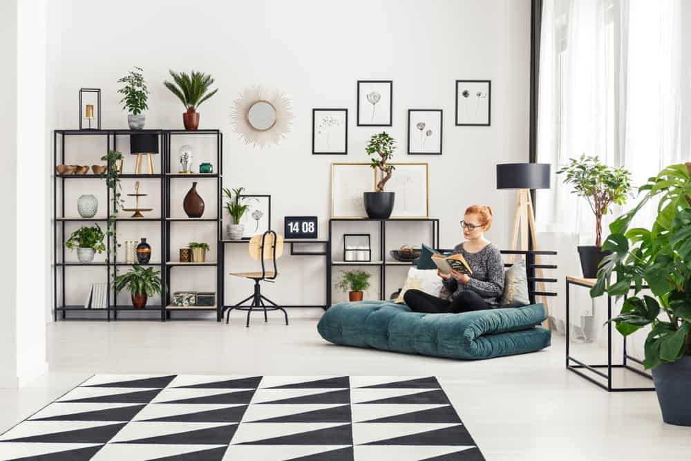 Girl in glasses sitting on a futon mattress and reading a book in bright interior with home office desk and fresh plants